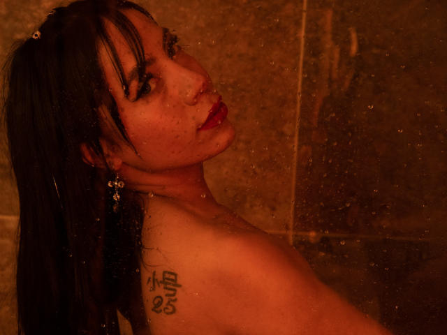 AmeliaRuso with wet black hair, red lipstick, and a tattoo on her right shoulder, looking back over her shoulder in a brown-tinted shower, water droplets on the glass.