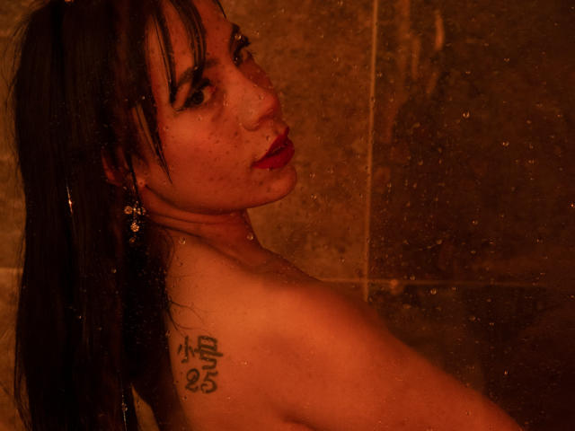 AmeliaRuso in a shower, wet black hair, red lipstick, tattoo on right shoulder, earrings, looking over shoulder, brown-tiled wall, water droplets on glass. Naked, medium breasts.