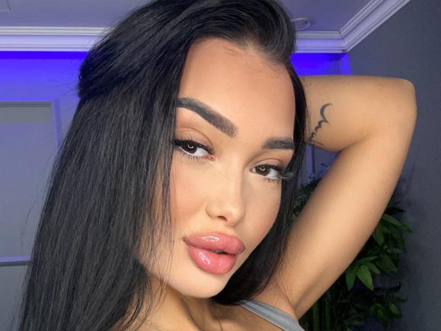 SashaFerrera with long black hair, brown eyes, full glossy pink lips, raised arm showing a small tattoo, and thick eyebrows, looking at the camera.