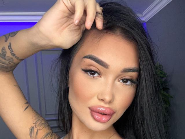 SashaFerrera with shiny pink lips, dark eyebrows, long black hair, tattooed arm, and brown eyes, holding hair up, looking at camera, in a selfie.