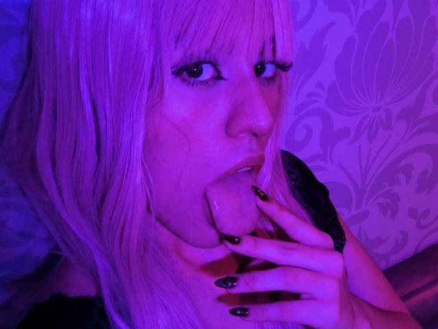 KaylinKrof with pink hair, black eyeliner, and black nails, sucking a large, pink, dildo-like object into her mouth, under purple lighting. Her nails are long and pointed.