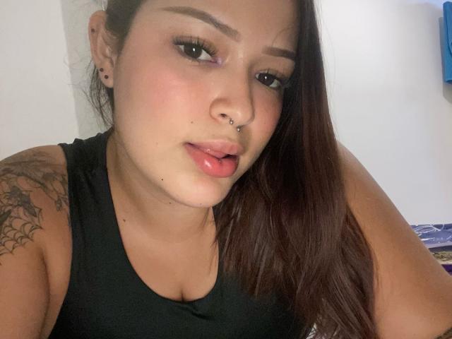 AllisonJustingb has brown hair, pink lipstick, and a nose piercing. She's wearing a black tank top, showing a tattoo on her left shoulder, and has a small black earring.
