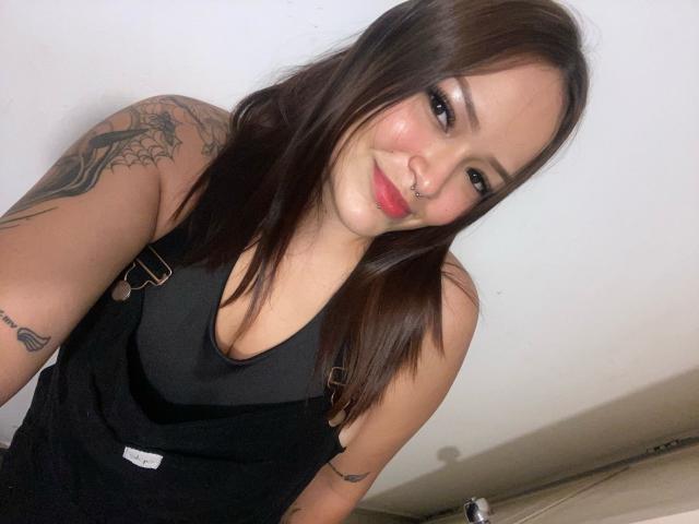 AllisonJustingb with brown hair, black tank top, nose ring, tattoos on arms, smiling, red lipstick, medium tits, light skin, white wall background, taking selfie.