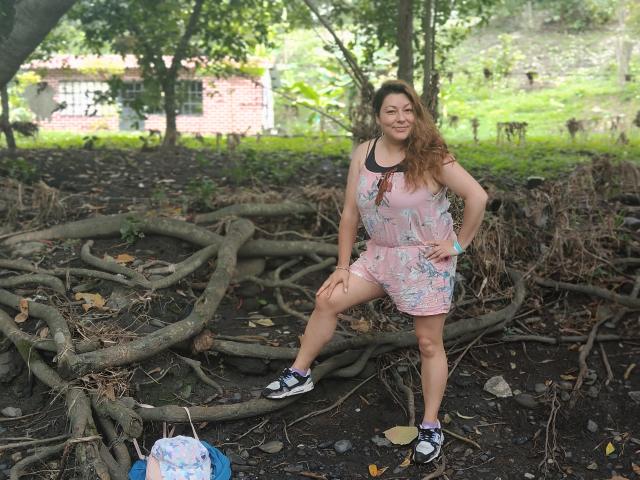 SakotoDika, de pie con una pierna levantada, vestida con un vestido floral rosa, zapatos negros y un collar, con cabello ondulado castaño, sonriendo en un área boscosa con raíces expuestas..