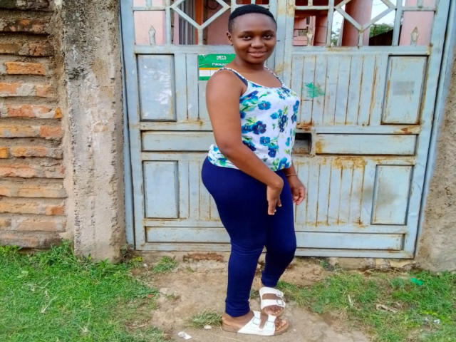 CuteJazmine has dark skin, a small bun, wearing a blue floral tank top, tight navy leggings, and white sandals, standing in front of a rustic blue gate.