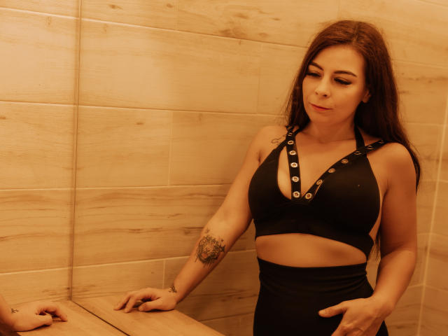 DiamondStar69, com cabelo castanho, sutiã preto com botões e calças pretas de cintura alta, tem uma tatuagem no braço direito, e está em pé em um cômodo com piso de madeira..
