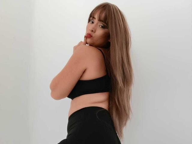 EmmaGoldyx, con cabello largo y castaño, un sostén negro y pantalones negros, mirando por encima del hombro con una expresión de puchero, mostrando su espalda desnuda y un atisbo de su trasero..