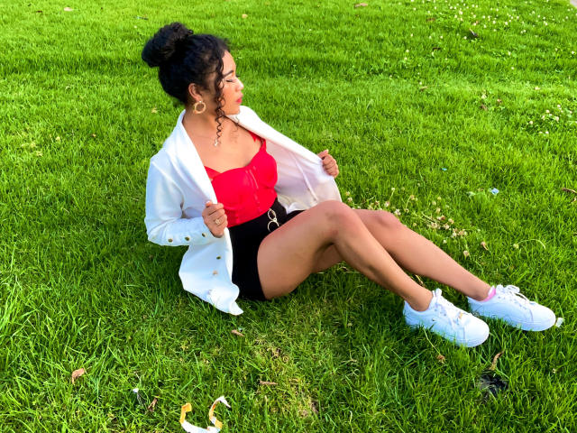 SophieSonyas sitting on grass, wearing a red top, black shorts, white jacket, white sneakers, curly hair in a bun, holding jacket edges, looking to the side, showing her tits.