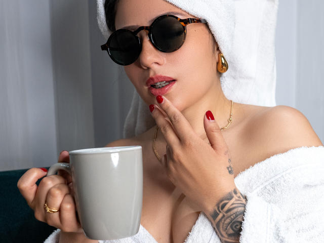 NahiaSan with a white towel on her head, wearing dark sunglasses, red lipstick, and a white robe, holding a gray mug, showing her red nails and tattooed hand.