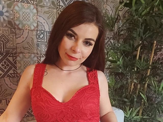 DiamondStar69 with red lace dress, showing tits, brown hair, red lipstick, black choker, and green eyes, standing next to a potted plant.