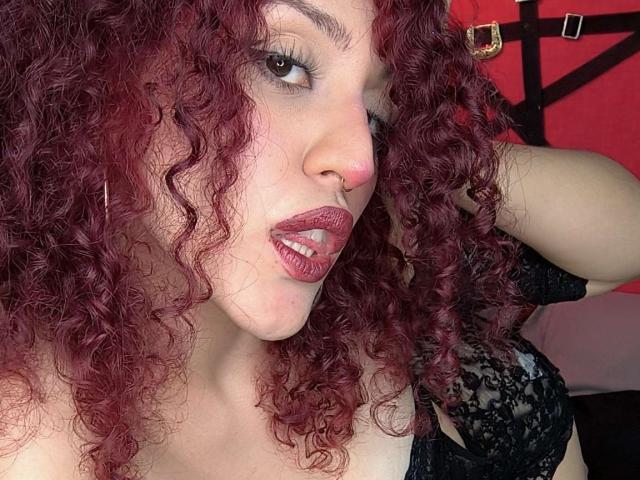 KaylinKrof with curly red hair, nose ring, and pink lipstick, looking at camera with tongue slightly out, wearing black lace top, showing her tits.