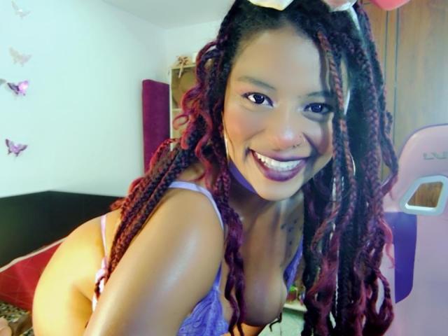 NashaJonny with dark skin and red-brown braids, smiling, wearing purple lace lingerie, showing her tits, nose ring, and purple lipstick, in a bedroom.