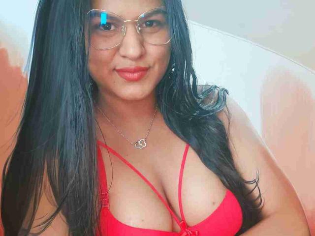 SweetAlma69 with long black hair, red bra, and heart necklace, wearing glasses, smiling, showing large tits, and wearing red lipstick.