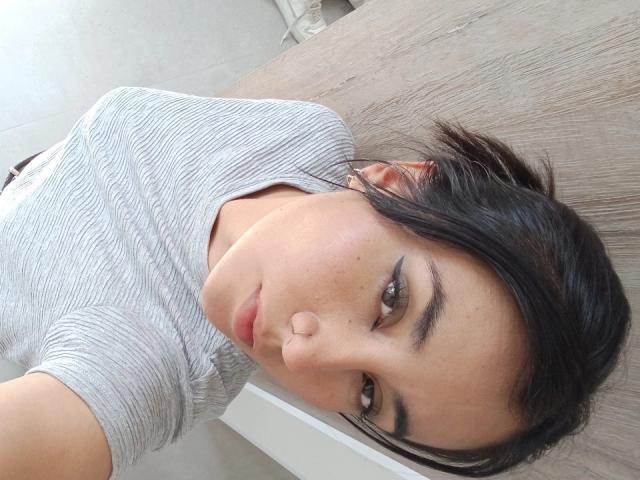 Natii69 has dark hair, brown eyes, and is wearing a gray striped shirt, lying on a wooden surface with a neutral expression. Her nose is slightly flattened.