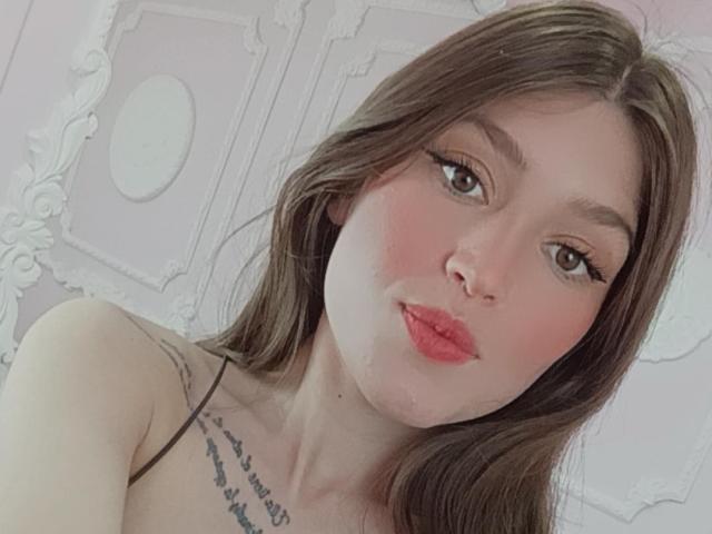 Tesakelle has pink lips, brown eyes, and long brown hair. She's pouting with a cute face, showing a tattoo on her shoulder. Her boobs are small, and she's topless.