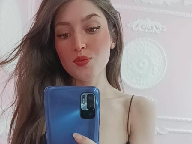 Tesakelle mit langen, braunen Haaren, voller, roter Lippen, die ein blaues Telefon hält, ihre kleinen Brüste zeigt und in einem Selfie eine niedliche Nase hat. Ihre Wimpern sind dicht und sie ist attraktiv..
