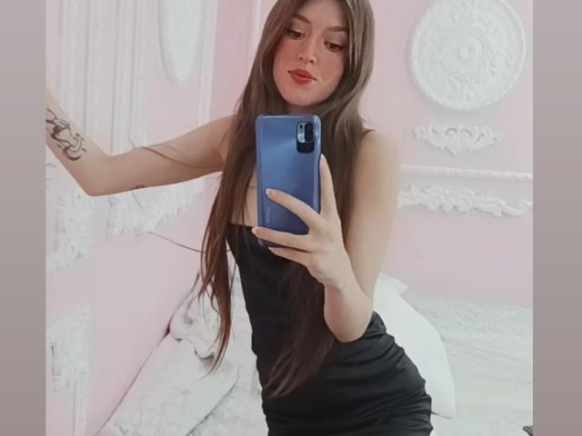 Tesakelle mit langen, braunen Haaren, die ein schwarzes Kleid trägt, macht ein Selfie mit ihrem blauen Handy, zeigt ein Tattoo auf ihrem Arm und trägt roten Lippenstift..