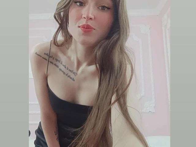 Tesakelle with long brown hair, black strapless top, pouty red lips, tattoo on right shoulder, and small tits, leaning forward, looking at camera.