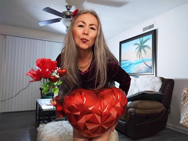 Eva Rodinson, com longos cabelos loiros, lábios carnudos, vestindo uma blusa escura de veludo, segurando um objeto vermelho brilhante em formato de coração, e um buquê de flores vermelhas em uma sala de estar..