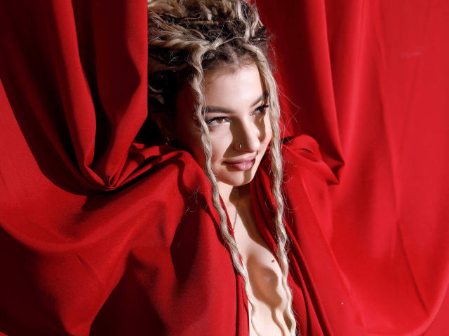 NoraSamuels with messy blonde braids, red cloth draped around, nose ring, smiling, cleavage visible, red background, soft light, slight shadow, confident, sexy, red dress partially open.