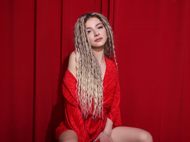 NoraSamuels with long blonde curls, red off-shoulder dress, sitting on red cloth, looking at camera, subtle smile, hands on thighs, cute, sexy, red background, soft light.
