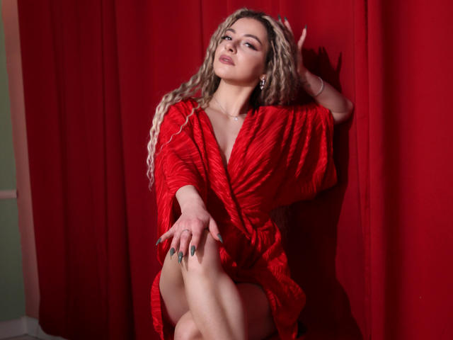 NoraSamuels with wavy blonde hair, red dress, and nipples slightly visible, sits against red curtain, right hand on head, left hand on knee, looking seductive.