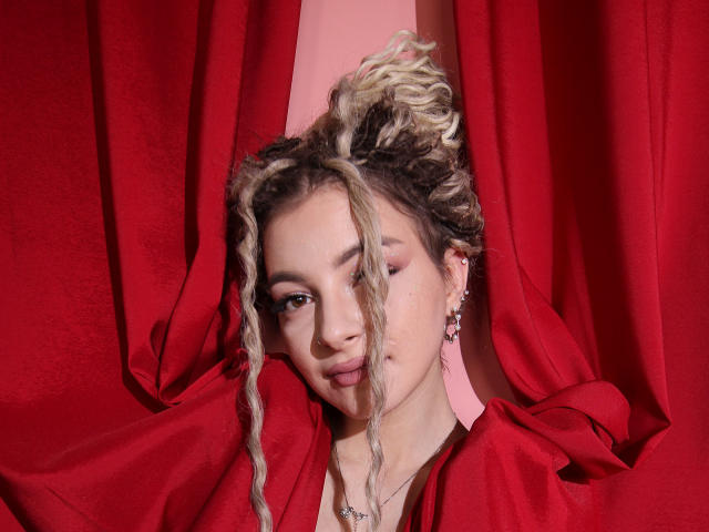 NoraSamuels with blonde and brown curly hair, pink eyeliner, and earrings, peeking through red curtains, looking slightly down, wearing a red cloth, necklace, and subtle makeup.