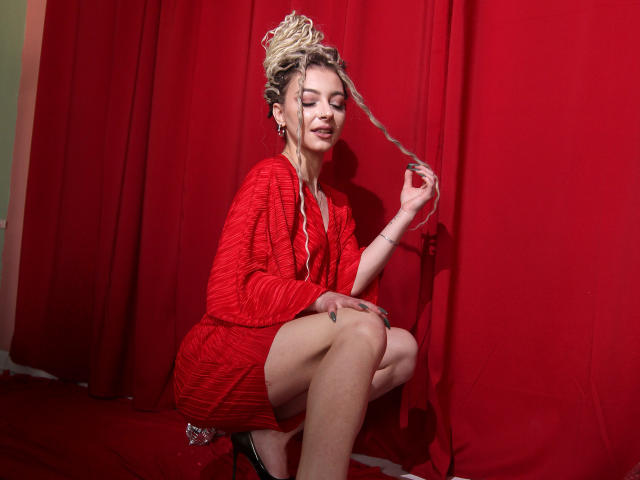NoraSamuels with blonde, curly updo, wearing red dress, squatting, holding hair, red background, smiling, red eyes, earrings, black high heels, red fabric behind her, right knee up.
