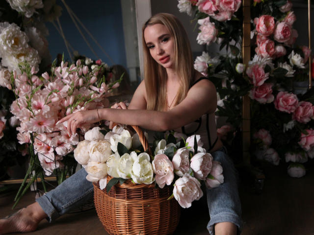 KiraSykes with long blonde hair, wearing a black top, sitting on the floor, holding a wicker basket filled with white and pink peonies, in front of a floral backdrop.