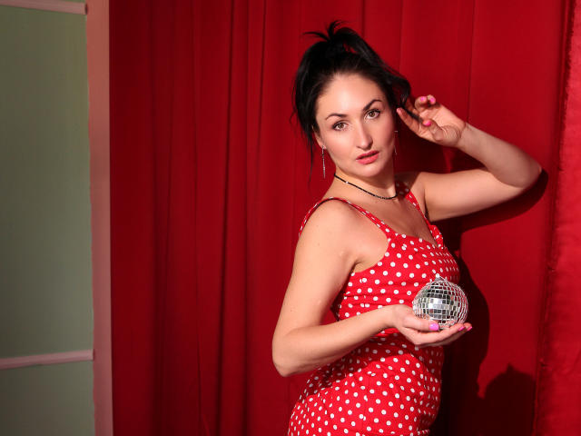 StefanaWinter with black hair in a ponytail, wearing a red polka dot dress, holding a silver disco ball, standing in front of red curtains, looking at the camera.