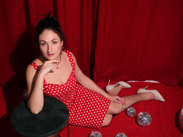 StefanaWinter with polka-dot red dress, sitting on red floor, white heels, hand on chin, balls around, dark hair in ponytail, green stool, red curtain, looking at camera.