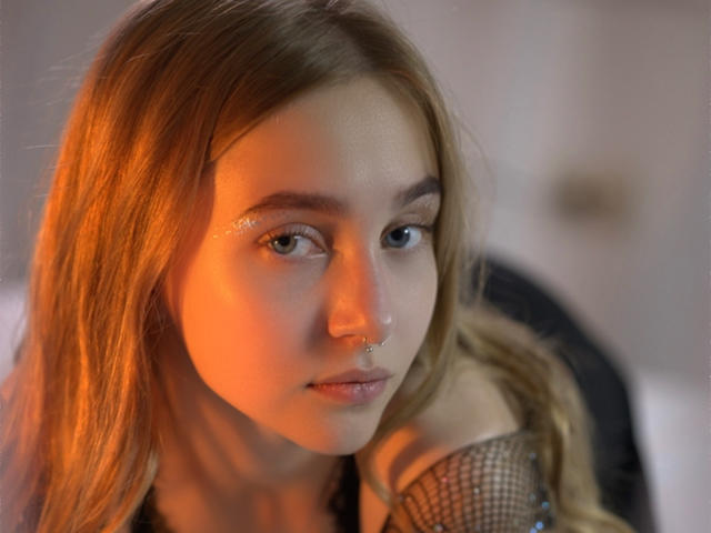 EmiliaMarshall has long brown hair, a nose ring, and sparkly eyelids. Her face is illuminated by orange light, and she's wearing fishnet sleeves. She looks serious.