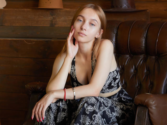 EmiliaMarshall with blonde hair, blue eyes, and small tits, wearing a black and white patterned dress, sitting on a brown leather chair, hand on cheek, looking at camera.