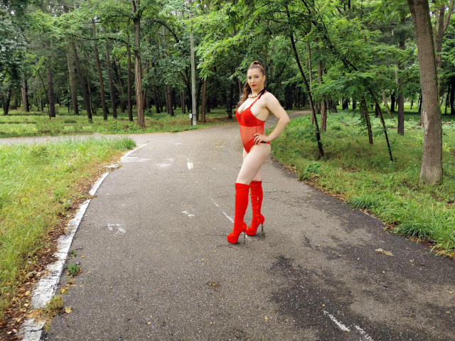 EmmaSeduction stands on a path in a forest, wearing a red one-piece swimsuit and matching knee-high boots, with her hand on her hip, showing her ass and tits.
