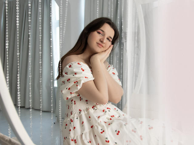 BonnieFlower with her hand on her cheek, wearing a white dress with red cherry patterns, sitting near hanging bead curtains, looking slightly to the side with a soft smile.