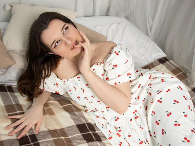 BonnieFlower with long brown hair, white cherry print dress, lying on bed, hand near mouth, looking at camera, white pillow, beige pillow, brown and white checkered blanket.