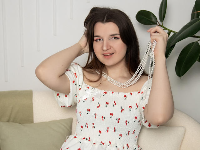 BonnieFlower with pale skin, brown hair, wearing a white dress with red floral patterns, holding a pearl necklace, sitting on a beige sofa, smiling, hand in hair.