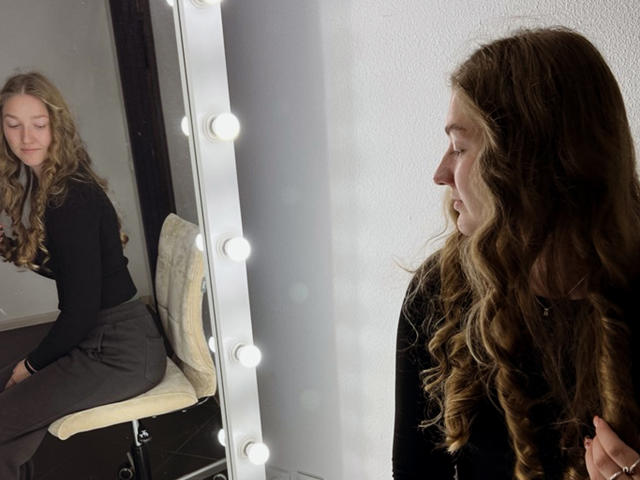 AgnesShorter with long wavy brown hair, black top, and gray pants, sitting on a chair in front of a mirror with lights, looking at her reflection.