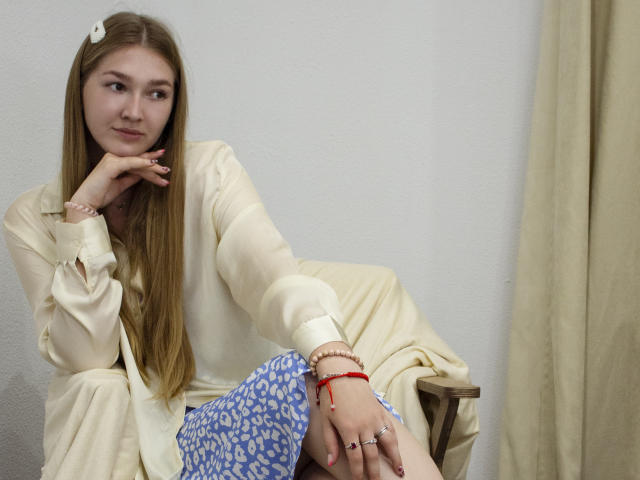 AgnesShorter with long blonde hair, white blouse, and blue patterned pants, sitting, hand on chin, looking to the side, wearing red bracelet and rings, beige curtain behind.