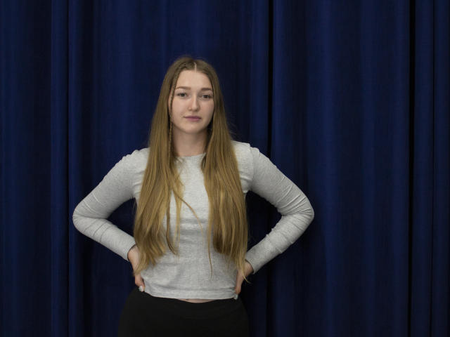 AgnesShorter with long blonde hair, grey long-sleeve shirt, black pants, hands on hips, standing against dark blue curtain, neutral expression, soft light, casual pose.