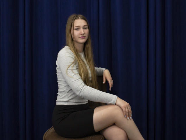 AgnesShorter with long blonde hair, gray long-sleeve shirt, black skirt, sitting on brown chair, crossed legs, blue curtain background, calm expression, subtle smile, fair skin.