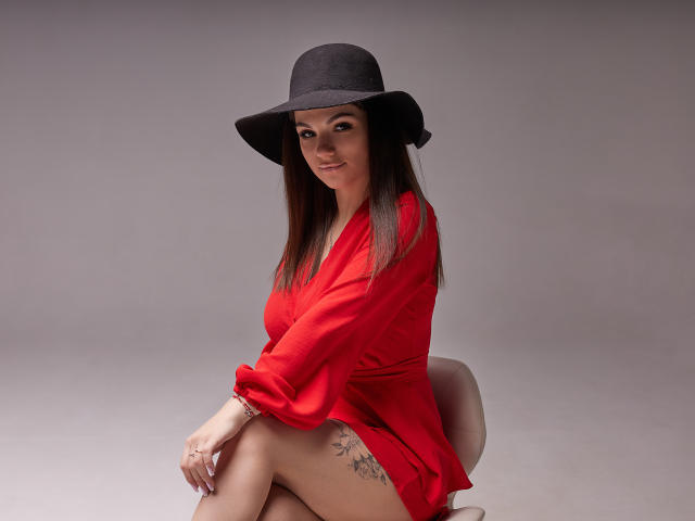 KarolinaSykes in a red dress, sitting with legs crossed, wearing a black hat, smiling, showing a tattoo on her right thigh, with long brown hair.