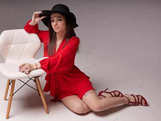 KarolinaSykes kneeling in red dress, black hat, red heels, white chair, hand on hat, tattoo on thigh, looking at camera, sexy, long brown hair, subtle smile.