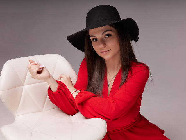 KarolinaSykes with long brown hair, wearing a black hat and red dress, sitting on a white chair, touching her pink-nailed fingers. Her tits are small, and she's smiling.