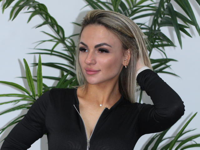 KarolinaSykes with blonde hair, wearing a black zip-up, has a small nose piercing, hand in hair, slight smile, green plant leaves in background, looking sexy and confident.