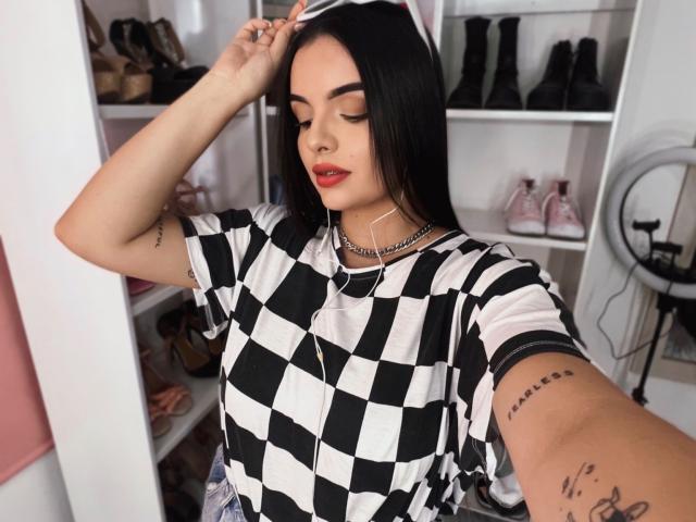 NovaDixon with black hair, wearing a black and white checkered shirt, red lipstick, earbuds, and tattoos, taking a selfie, showing off her tits and ass in a room.