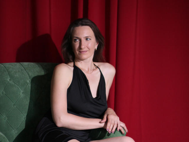 AngelicaShackley with brown hair, wearing a black halter dress, sitting on a green velvet couch, red curtain background, smiling slightly, hands resting on her thigh.