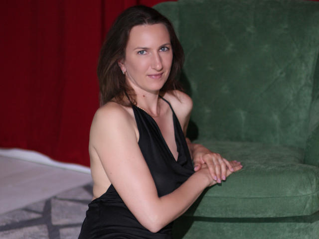 AngelicaShackley in a black dress with a deep neckline, sitting on a green velvet couch, smiling with hands on the couch, brown hair, blue eyes, no visible boobs.