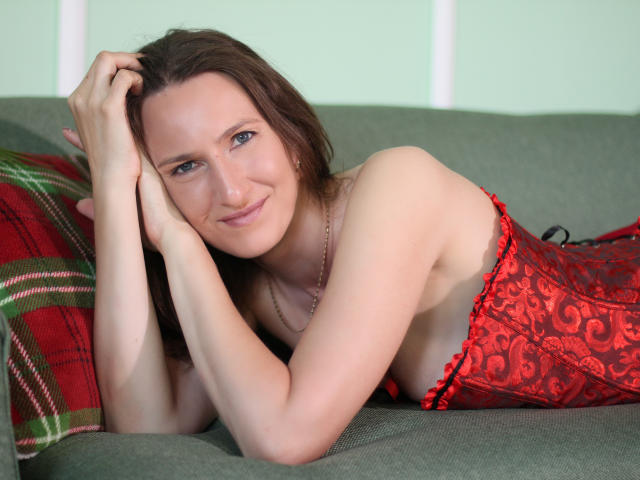 AngelicaShackley lying on a green couch, wearing a red lace corset, smiling with brown hair, hand on her head, showing her tits and a cute freckle on her cheek.