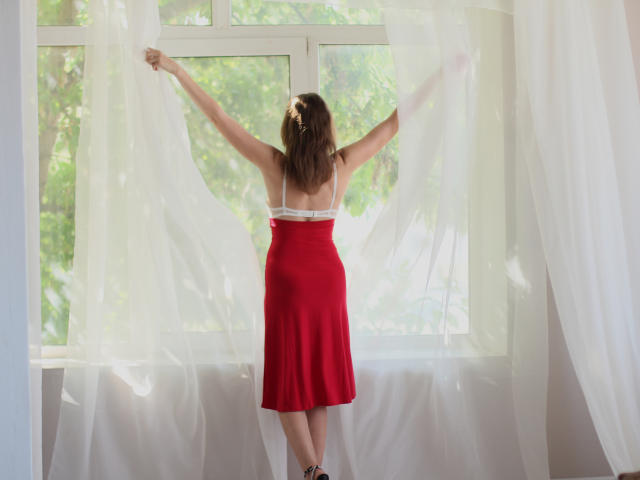 AngelicaShackley stands with arms raised, white curtains in front of a window, wearing a red dress with white straps, brown hair, and black shoes.